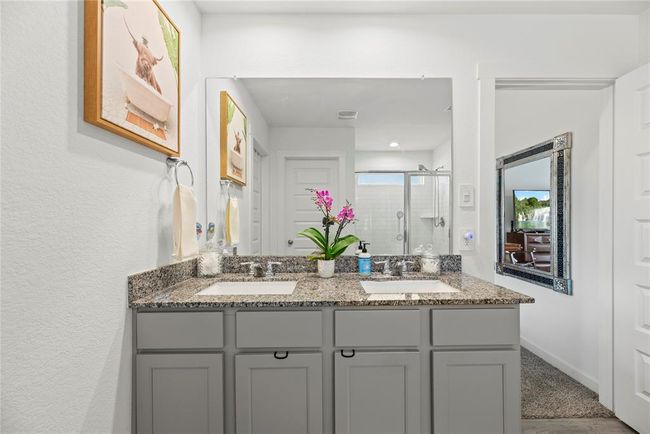 The primary bathroom vanity is designed with two square sinks, each equipped with stylish nickel faucets. The gray cabinets offer a modern, sleek look, while the light natural wood-colored LVP flooring adds warmth and a touch of nature to the space. This combination of finishes creates a sophisticated yet inviting atmosphere, balancing contemporary design with a touch of earthy charm. The polished nickel details complement the overall aesthetic, making the bathroom both functional and visually appealing. | Image 13