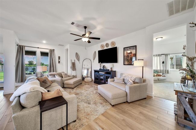 Bright and spacious living room with hardwood floors, neutral tones, and ample seating. Features include a ceiling fan, large TV, and decorative accents. Large windows and glass doors offer natural light and a view of the backyard. Adjacent room visible, enhancing the open layout. | Image 29