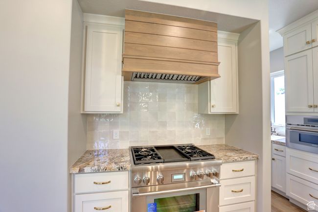 Kitchen with appliances with stainless steel finishes, custom exhaust hood, tasteful backsplash, white cabinetry, and light stone counters | Image 9