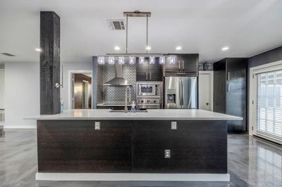 Kitchen featuring dark brown cabinets, stainless steel appliances, an island with sink, wall chimney exhaust hood, and sink | Image 1