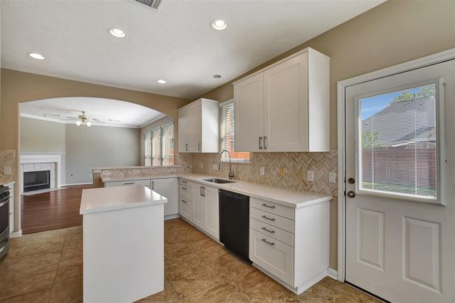 This photo showcases a bright, modern kitchen with white cabinetry, a central island, and tiled flooring. It opens into a living area with hardwood floors and a fireplace. Ample natural light comes through large windows, and there's a door leading to the outdoor space. | Image 24