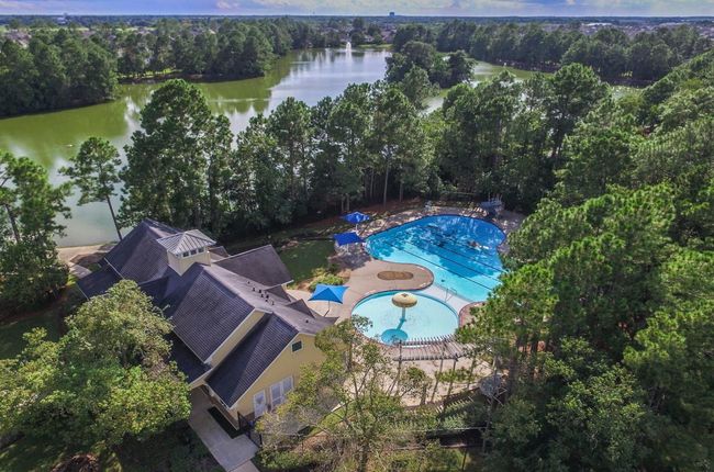 This photo showcases the clubhouse featuring a large pool area with umbrellas and splash pad surrounded by lush trees, and adjacent to a picturesque lake, offering a tranquil and private setting. | Image 37