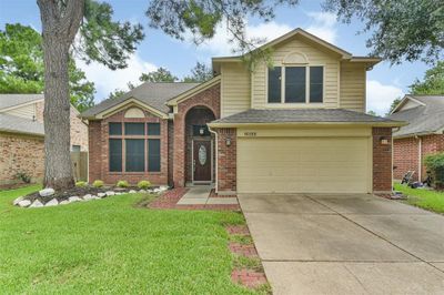 This is a two-story, single-family home featuring a brick facade with contrasting upper siding, a two-car garage, and a well-maintained front lawn with a mature tree. The entryway is accented with an arched detail above the front door. | Image 1