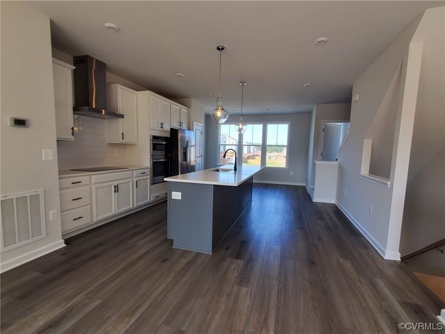 Kitchen featuring dark hardwood floors, kitchen island with sink, backsplash, white cabinets, wall chimney range hood, light countertops, appliances with stainless steel finishes, and pendant lighting | Image 18