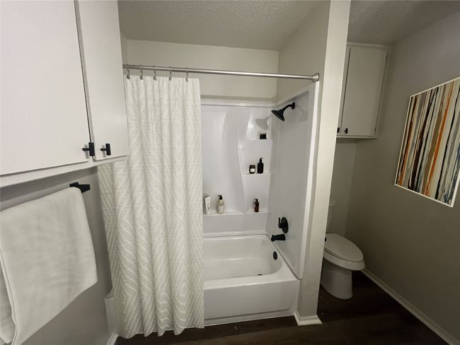 Bathroom with a textured ceiling, toilet, wood finished floors, shower / bath combo with shower curtain, and baseboards | Image 5