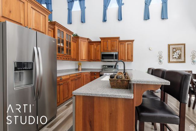 Kitchen featuring stainless steel appliances, a towering ceiling, brown cabinets, light countertops, and an island with sink | Image 11