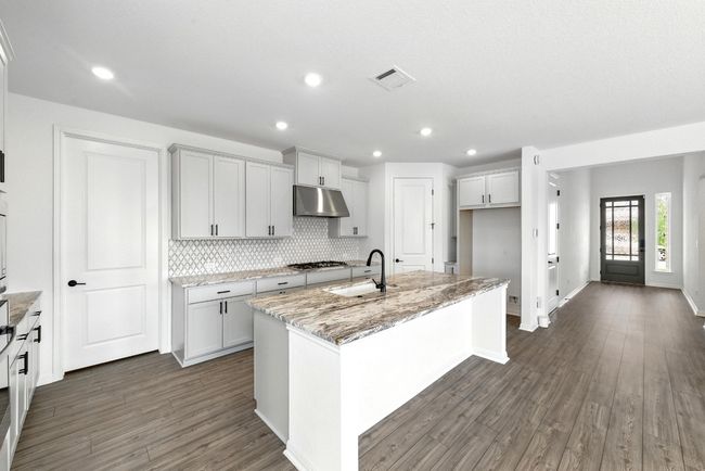 Kitchen with under cabinet range hood, a center island with sink, backsplash, light stone counters, and a sink | Image 11