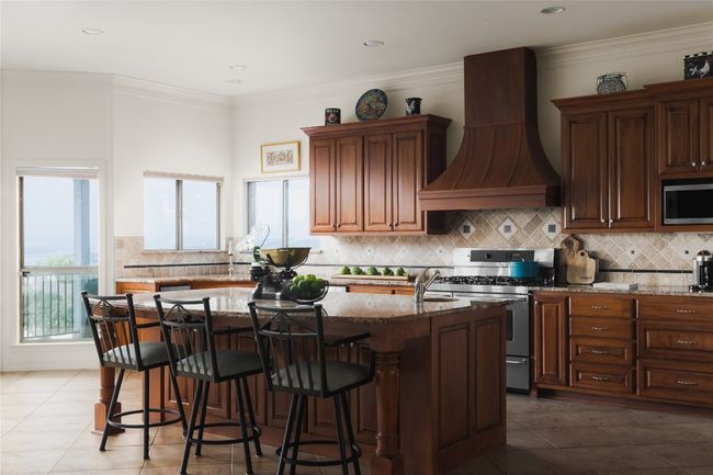 Kitchen with custom range hood, stainless steel appliances, light stone countertops, a kitchen island with sink, and backsplash | Image 19