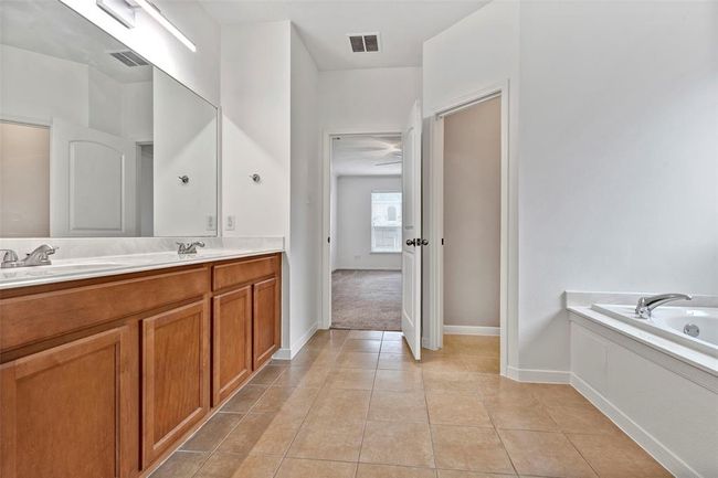 Spacious bathroom with double sinks, large mirror, and wooden cabinetry. Features a bathtub and connects to a carpeted bedroom, providing a bright and functional space. | Image 16