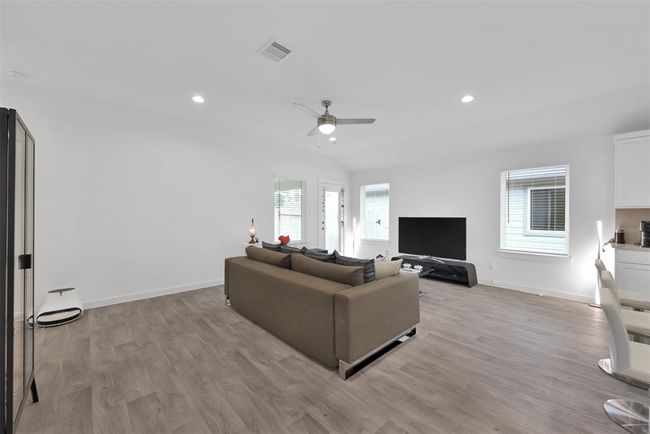 This spacious living room features a modern, open layout with light wood flooring and ample natural light. It includes a comfortable seating area, ceiling fan, large TV, and easy access to the outdoors, creating a welcoming and functional space. | Image 10