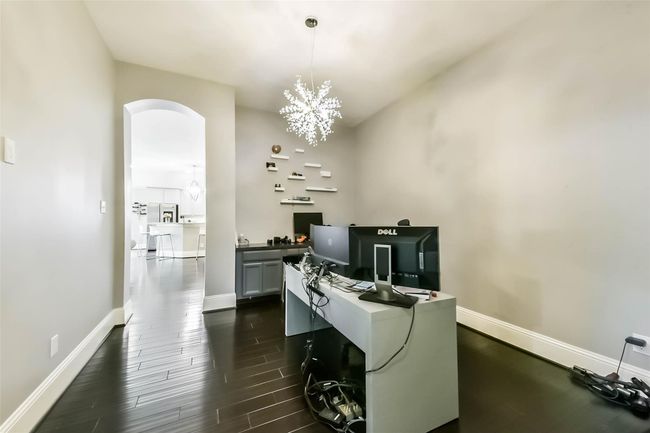 A modern home office with dark hardwood floors and a unique chandelier. The room opens to a bright kitchen area through an archway, creating an open and connected feel. | Image 21
