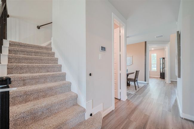 This photo showcases a bright, inviting hallway with a carpeted staircase leading to the upper floor. The hallway features wood-style flooring and leads to multiple rooms, offering a glimpse of a cozy, well-lit space with plenty of natural light. | Image 10