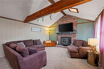 Living room with light colored carpet, a wood stove, track lighting, wooden walls, and lofted ceiling with beams | Image 3