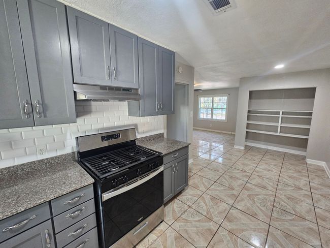 Kitchen with stainless steel gas range, under cabinet range hood, gray cabinetry, backsplash, and dark stone counters | Image 26