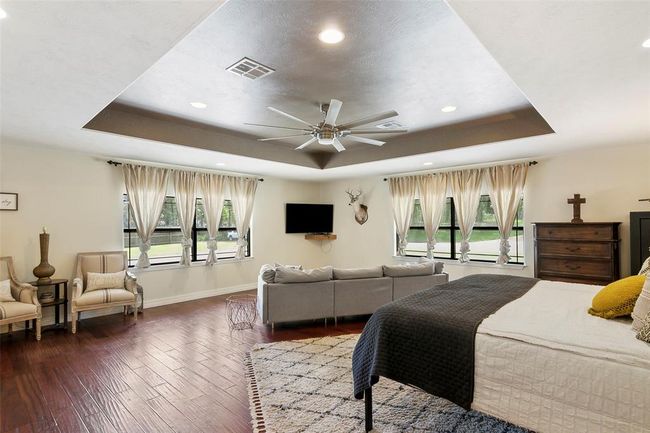 Spacious master bedroom with tray ceiling, hardwood floors, and abundant natural light from large windows. Features a cozy seating area, mounted TV, and elegant furnishings, creating a comfortable and inviting atmosphere. | Image 27