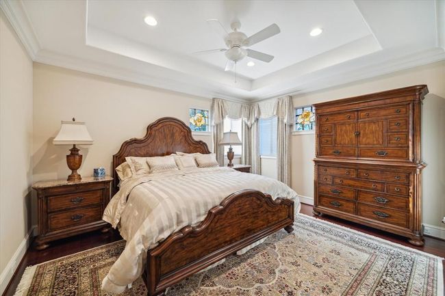 This elegant bedroom features a tray ceiling with recessed lighting and a ceiling fan. The room is accented by stained glass windows and a plush area rug, creating a warm and inviting atmosphere. | Image 21