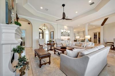 This is a spacious, elegantly appointed living room featuring high ceilings, a classic fireplace, large windows for natural light, and an open-concept layout leading into a highly-functional kitchen. | Image 3