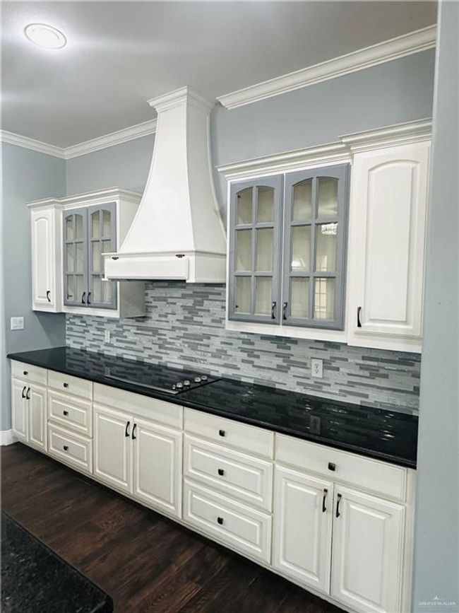 Kitchen with black electric cooktop, custom exhaust hood, dark countertops, ornamental molding, and backsplash | Image 5