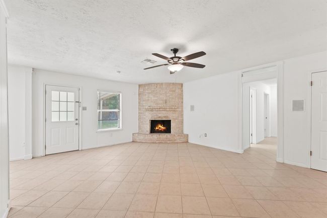This spacious living room features a cozy brick fireplace, neutral tile flooring, and a ceiling fan. It has plenty of natural light through a window and a door leading outside, creating a bright and welcoming atmosphere. | Image 9