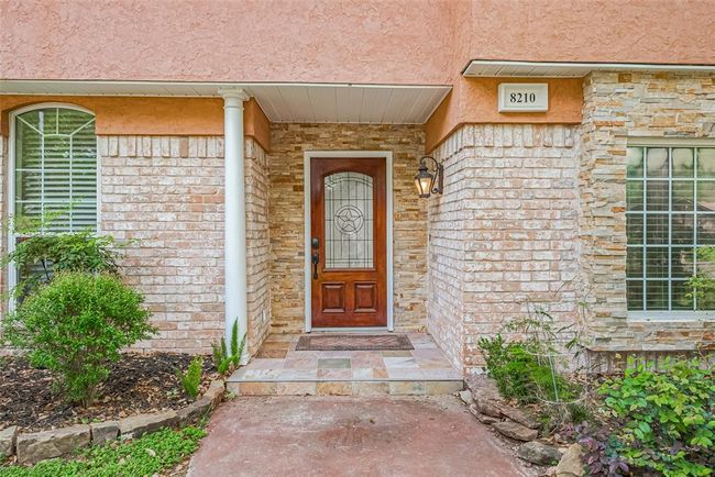 The inviting front porch features stacked stone, tile flooring, and a gorgeous wood door with decorative glass inlay. A classic lantern-style sconce sets the tone for the warmth and character found throughout the home. | Image 4