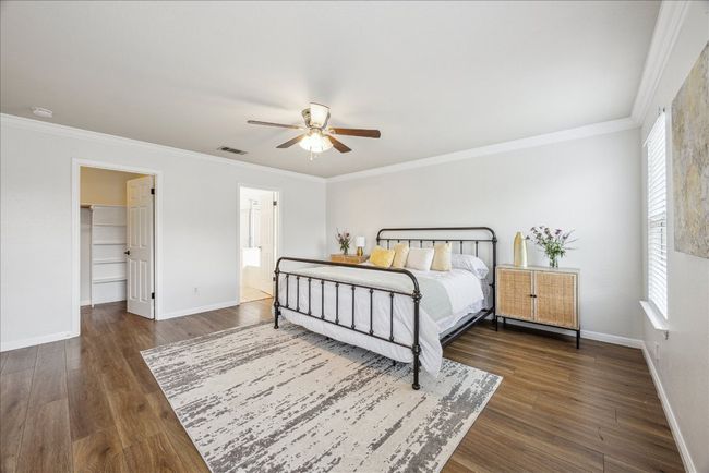 Primary bedroom with view of walk-in closet and bathroom, featuring crown molding, vinyl flooring, large windows allowing natural light from the yard, and a ceiling fan | Image 14