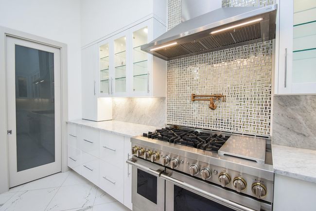 This modern kitchen features sleek white cabinetry, a stainless steel gas range with a striking feature backsplash, and a Carrarra marble countertop and backsplash. It offers ample storage and a contemporary design, ideal for culinary enthusiasts. | Image 19