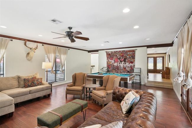 This spacious living room features hardwood floors, comfortable seating, and stylish decor. It includes a pool table for entertainment, large windows for natural light, and a ceiling fan. The room is accented with a decorative wall hanging and a mounted deer head, creating a cozy and inviting atmosphere. | Image 11