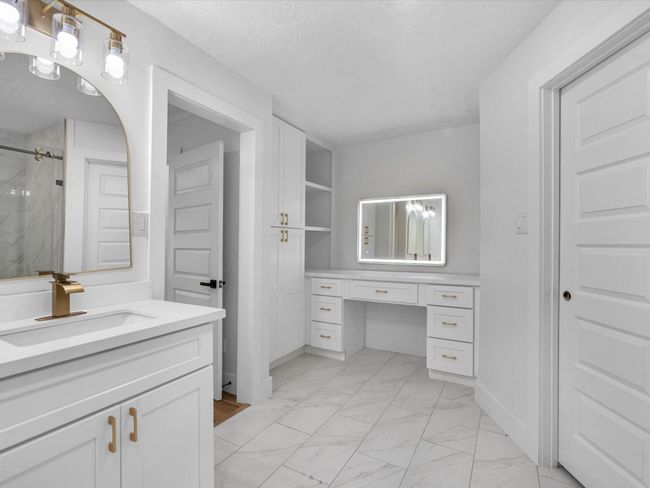 This bathroom features a modern design with white cabinetry, gold hardware, and a sleek marble-like tile floor. It includes a spacious vanity with a large illuminated mirror, ample storage, and elegant lighting fixtures for a bright and airy feel. | Image 24