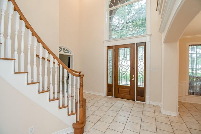 This welcoming foyer features a grand staircase with wooden handrails and white spindles, a tiled floor, and elegant double doors with decorative glass panels, highlighted by a large arched window above. | Image 9