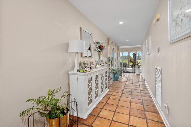 A long, inviting hallway with warm-toned tile flooring leads into the home, offering a seamless flow toward the main living spaces. Neutral walls and recessed lighting create a bright and airy feel, while large windows at the far end frame stunning views of the outdoors. | Image 8