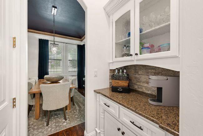 The butler pantry nestled between the kitchen and formal dining rooms features white cabinets with glass inserts, tasteful stone backsplash, granite countertop, decorative hardware and wood floors | Image 14