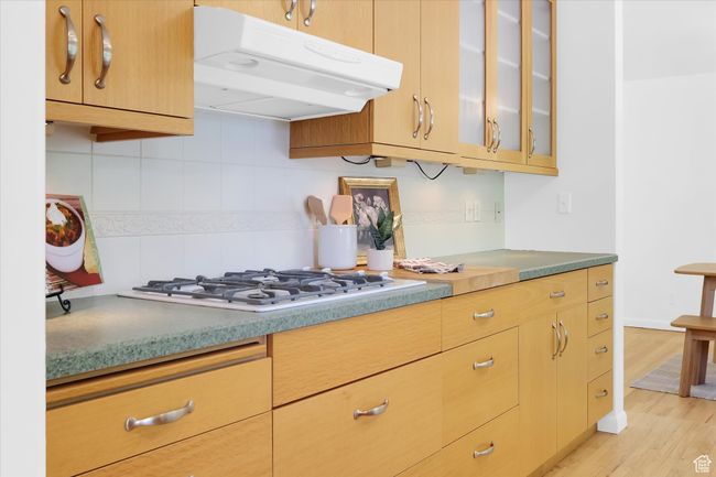 Kitchen with under cabinet range hood, white gas cooktop, decorative backsplash, glass insert cabinets, and light wood-type flooring | Image 10