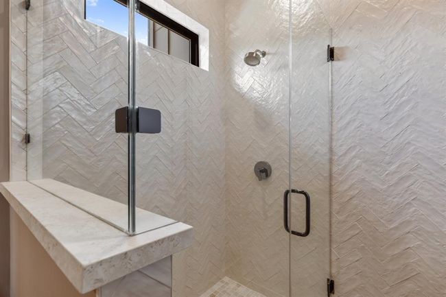 Pearlescent subway tile have been set in a herringbone pattern to add texture and drama. The shower enclosure features frameless glass and a curb, bench and ledge customized from Consentino porcelain slabs. A window offers natural light while maintaining privacy. | Image 25