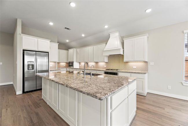 This stunning kitchen is the heart of the home, showcasing an oversized granite island with a deep basin sink, on-demand hot water, and bar seating for effortless prep and casual dining. A custom hood vent adds a stylish focal point above the cooktop, while stainless steel appliances, a herringbone tile backsplash, under-cabinet lighting, and extensive cabinetry offer the perfect blend of beauty and functionality. | Image 16