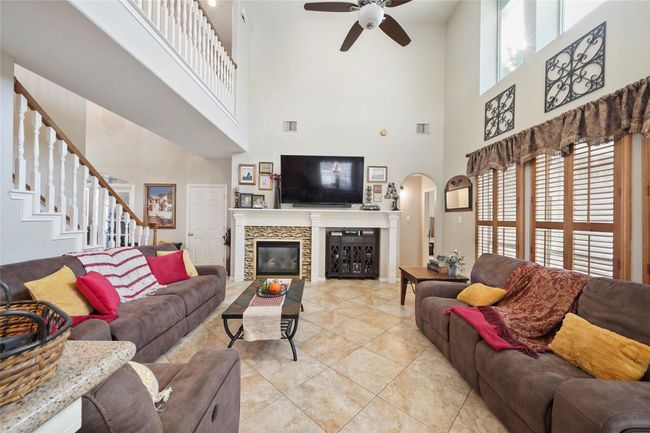 This spacious living room boasts high ceilings and abundant natural light. It features a cozy fireplace, large windows with wooden shutters, and elegant tile flooring. The open layout includes a staircase leading to the upper level, adding a touch of grandeur. Perfect for comfortable living and entertaining. | Image 15