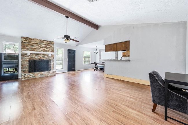 This spacious living area features a vaulted ceiling with a wooden beam, hardwood floors, and a stone fireplace. It offers an open layout with a view into the kitchen, a ceiling fan, and abundant natural light from large windows and a glass door. | Image 6