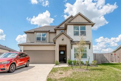 This is a modern two-story home featuring a neutral color palette with a mix of siding and stone facade. It includes an attached two-car garage, a well-maintained lawn, and a young tree in the front yard, under a clear blue sky. | Image 1