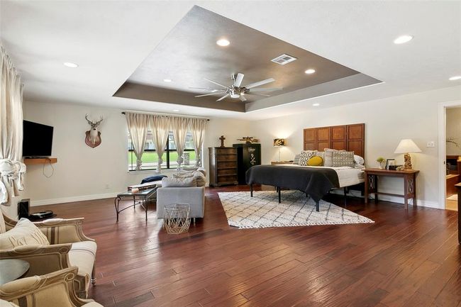 This spacious bedroom features elegant hardwood floors, a coffered ceiling with recessed lighting, and ample natural light from multiple windows. It includes a cozy seating area, stylish decor, and a mounted TV, offering both comfort and functionality. | Image 24