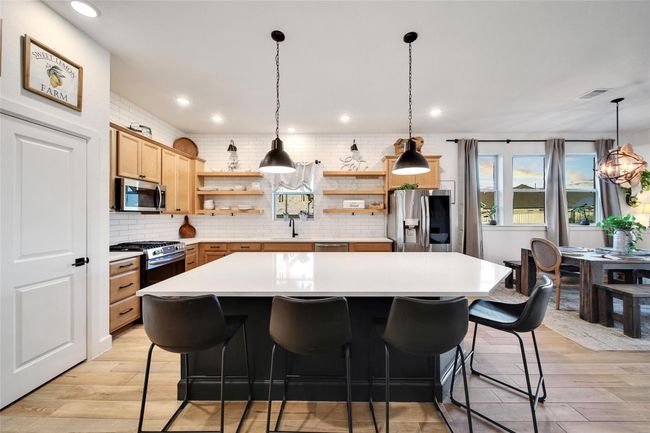 This kitchen features a modern farmhouse design with a large central island, sleek black barstools, and pendant lighting. It has warm wood cabinetry, stainless steel appliances, and open shelving. Adjacent is a cozy dining area with ample natural light from large windows, creating an inviting atmosphere. | Image 18