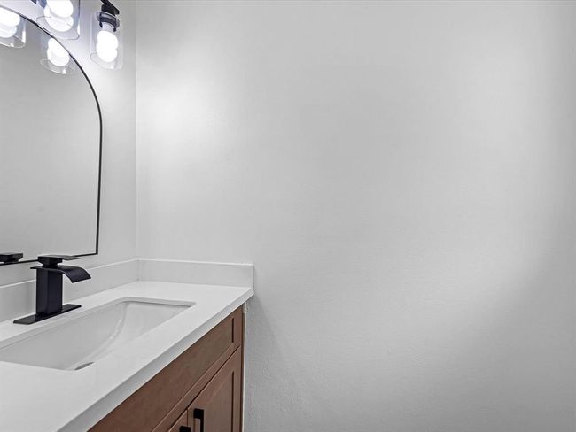 This photo shows a modern bathroom with a sleek white countertop and a rectangular sink. The vanity features a dark faucet and wood cabinetry, complemented by a large, stylish mirror with contemporary lighting above. The overall look is clean and minimalistic. | Image 18