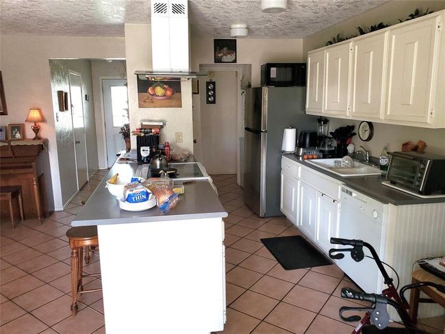 This kitchen features white cabinetry, a central island with seating, and modern appliances including a stainless steel fridge. It's spacious with tiled flooring and has ample counter space, ideal for meal prep and entertaining. | Image 5