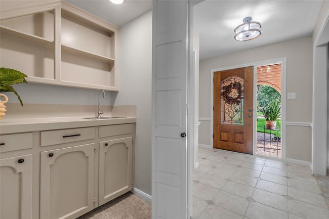 The photo shows an inviting entryway with a wooden door featuring decorative glass, opening to a well-lit space with tile flooring. Adjacent is a built-in wet bar with open shelving, offering functional storage and utility. | Image 13