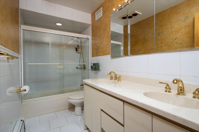 Bathroom featuring a baseboard heating unit, double vanity, bath / shower combo with glass door, tile patterned floors, and tile walls | Image 9