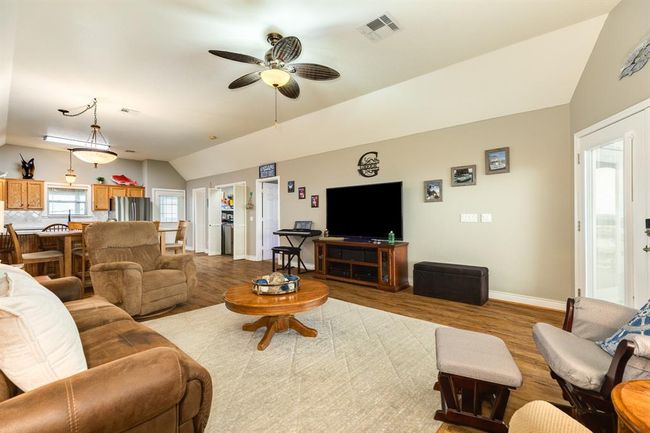 This spacious open-concept living area features a cozy sitting space with plush seating, a large flat-screen TV, and a ceiling fan. The room seamlessly flows into a modern kitchen with wooden cabinets, stainless steel appliances, and a dining space, all highlighted by warm wood flooring. Perfect for family gatherings and entertaining. | Image 6