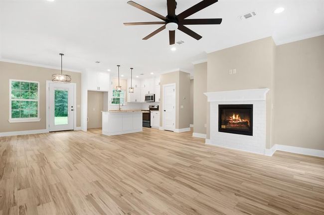 This photo showcases a spacious open-concept living area with light wood flooring and neutral walls. The room features a modern ceiling fan, a cozy fireplace, and ample natural light from large windows. The kitchen is equipped with stainless steel appliances, white cabinetry, and a central island with pendant lighting. A door leads to an outdoor space, enhancing the home's airy feel. | Image 5