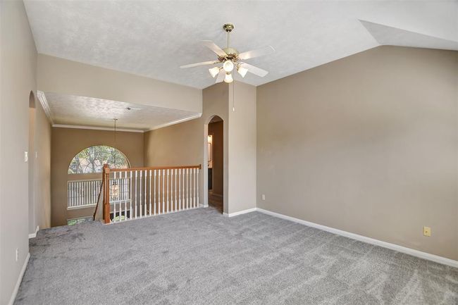This room features a cozy, carpeted area with neutral walls, a ceiling fan, and an arched window. It's located on an upper floor with a railing overlooking the staircase, creating an open and inviting space. | Image 29