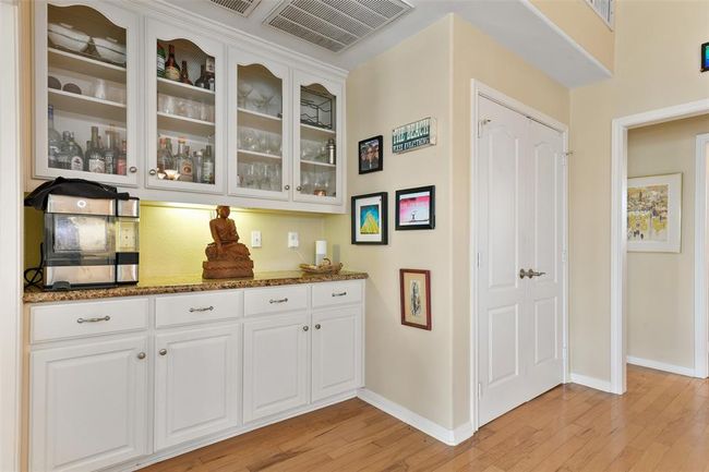 This photo shows a cozy hallway area featuring built-in white cabinetry with glass doors, perfect for displaying glassware and bottles. The space includes a small countertop with decorative items, warm bamboo wood flooring, and a few framed artworks on the walls, adding a personal touch. | Image 20