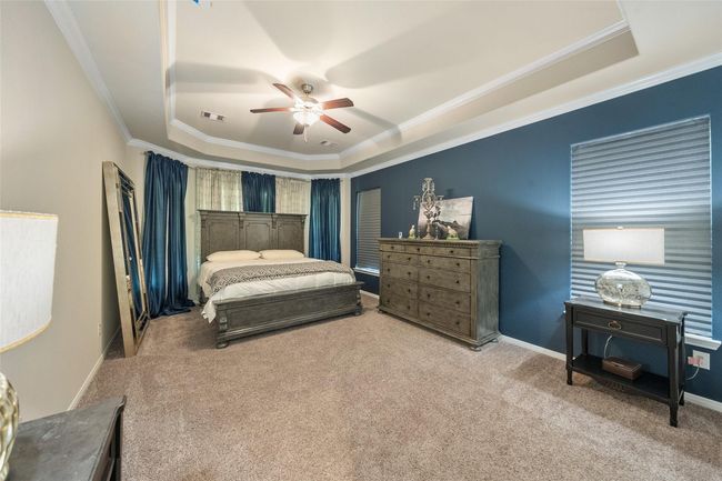 This spacious primary bedroom features a tray ceiling, neutral carpet, and a bold blue accent wall. It includes a bay window with ample natural light. | Image 16