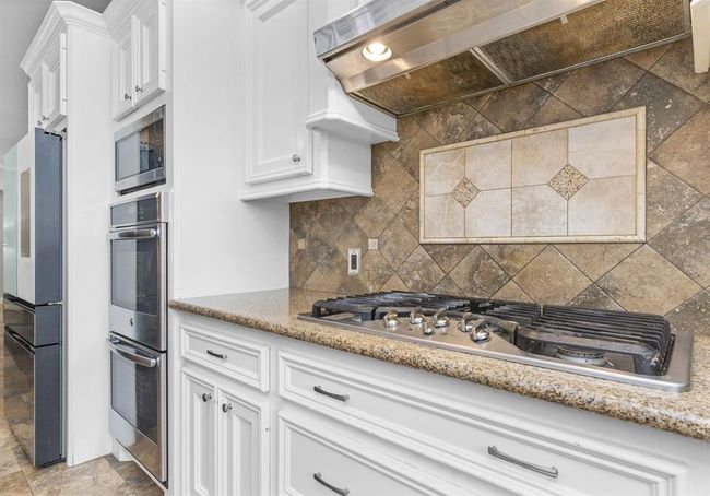 This kitchen features white cabinetry, stainless steel appliances including a wall oven and microwave, a gas cooktop, and granite countertops with a decorative tile backsplash. | Image 23