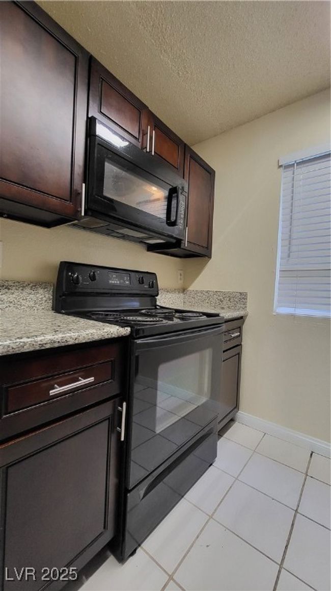 Kitchen with light tile patterned flooring, black / electric stove, a textured ceiling, and dark brown cabinets | Image 5